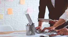 wo people leaning on desk with computer and one person holding a tablet